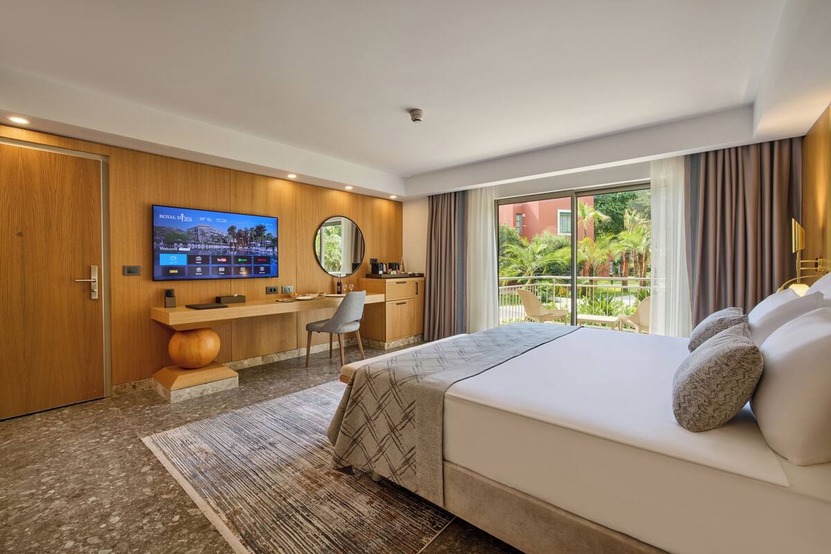A modern hotel room with a large bed, a desk, a flat-screen TV, and a balcony with a view of greenery.
