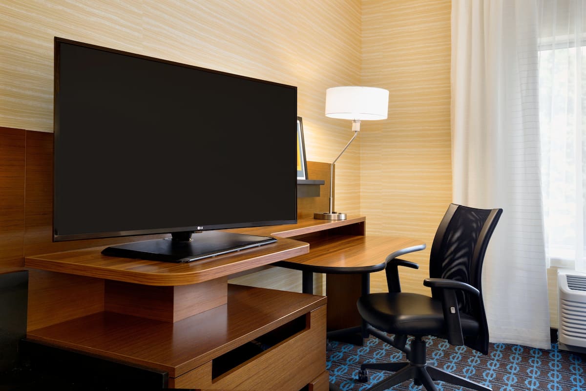 A hotel room with a flat-screen TV on a wooden desk, a black office chair, a floor lamp, and a window with sheer curtains.
