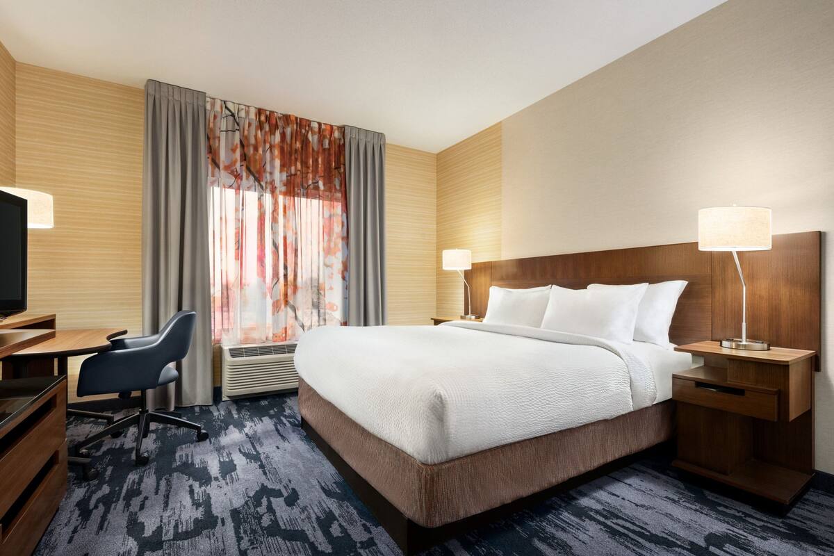 Ein Hotelzimmer mit einem großen Bett, einem Schreibtisch mit Stuhl, einem Fernseher und einem Fenster mit Vorhängen.