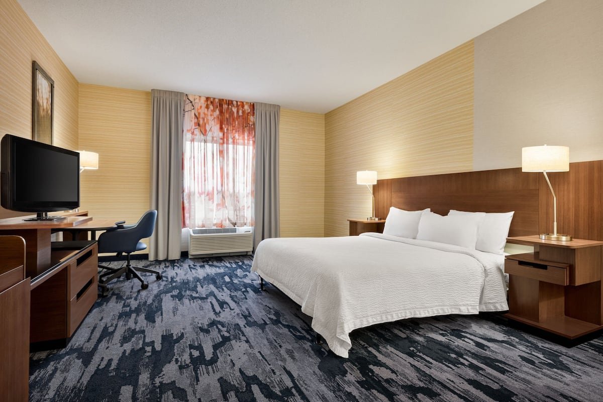 Ein Hotelzimmer mit einem großen Bett, einem Schreibtisch mit Computer, einem Fernseher und einem Fenster mit Vorhängen.