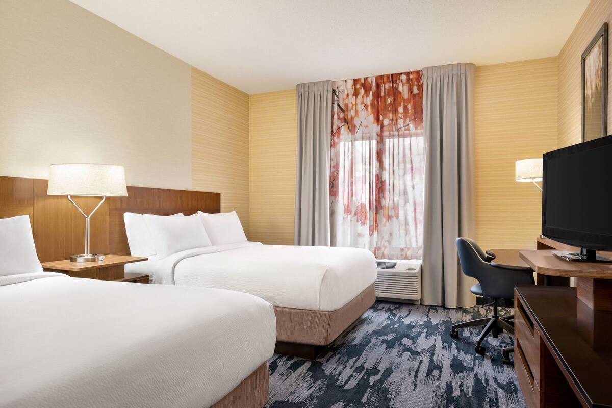 Ein Hotelzimmer mit zwei Betten, einem Schreibtisch, einem Fernseher und einem Fenster mit Vorhängen.