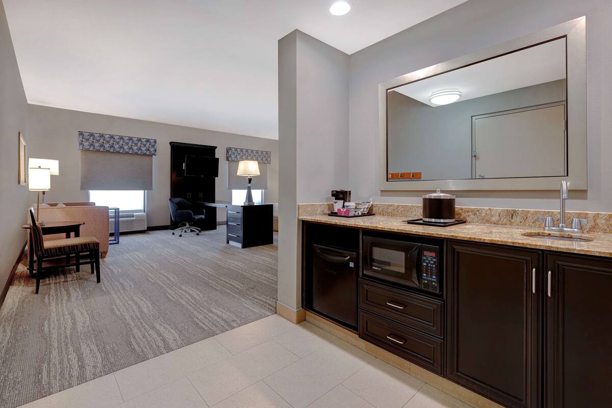 A hotel room with a kitchenette, a large mirror, a dining area with a table and chairs, and a carpeted floor.