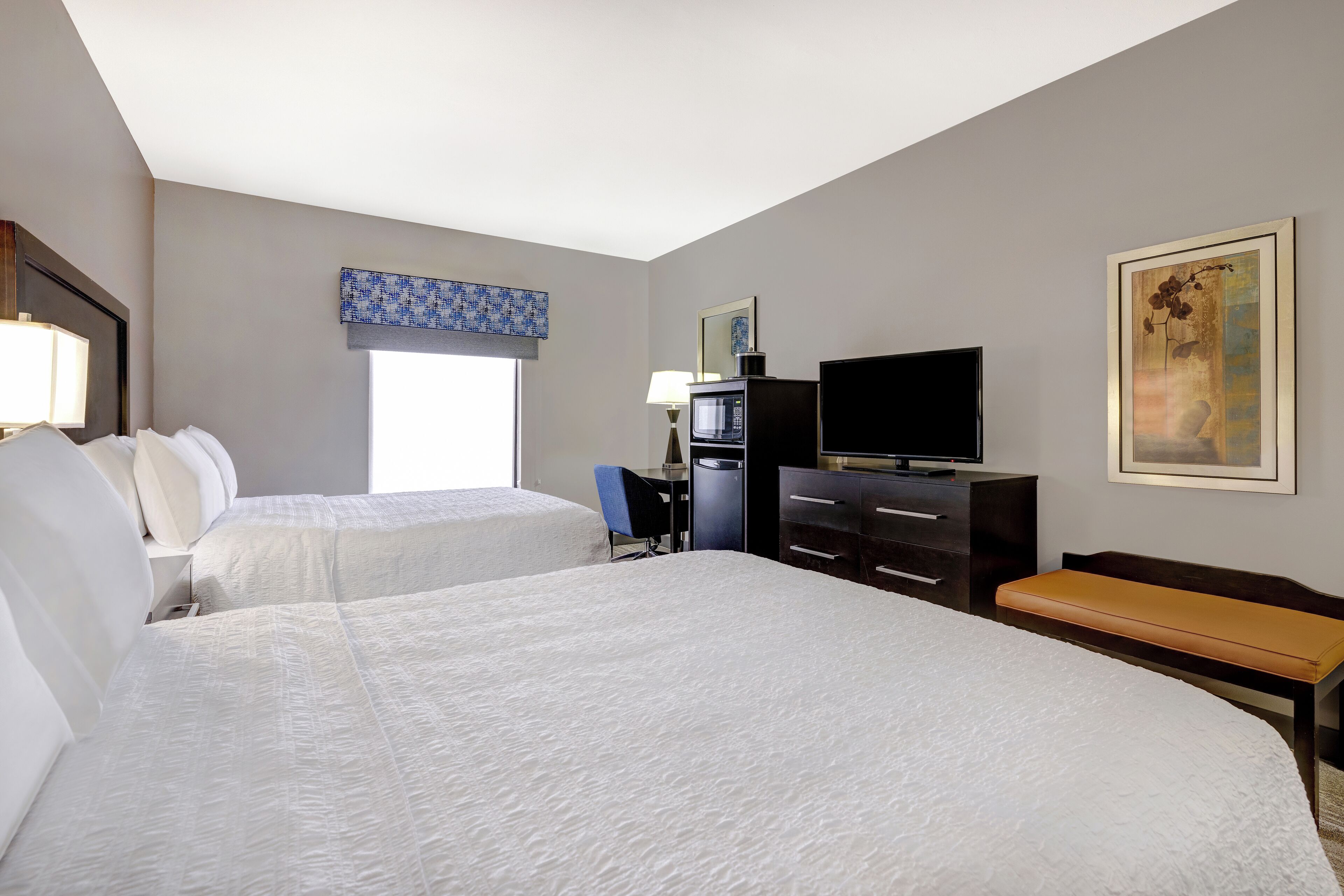 A hotel room with two beds, a desk, a television, and a painting on the wall.