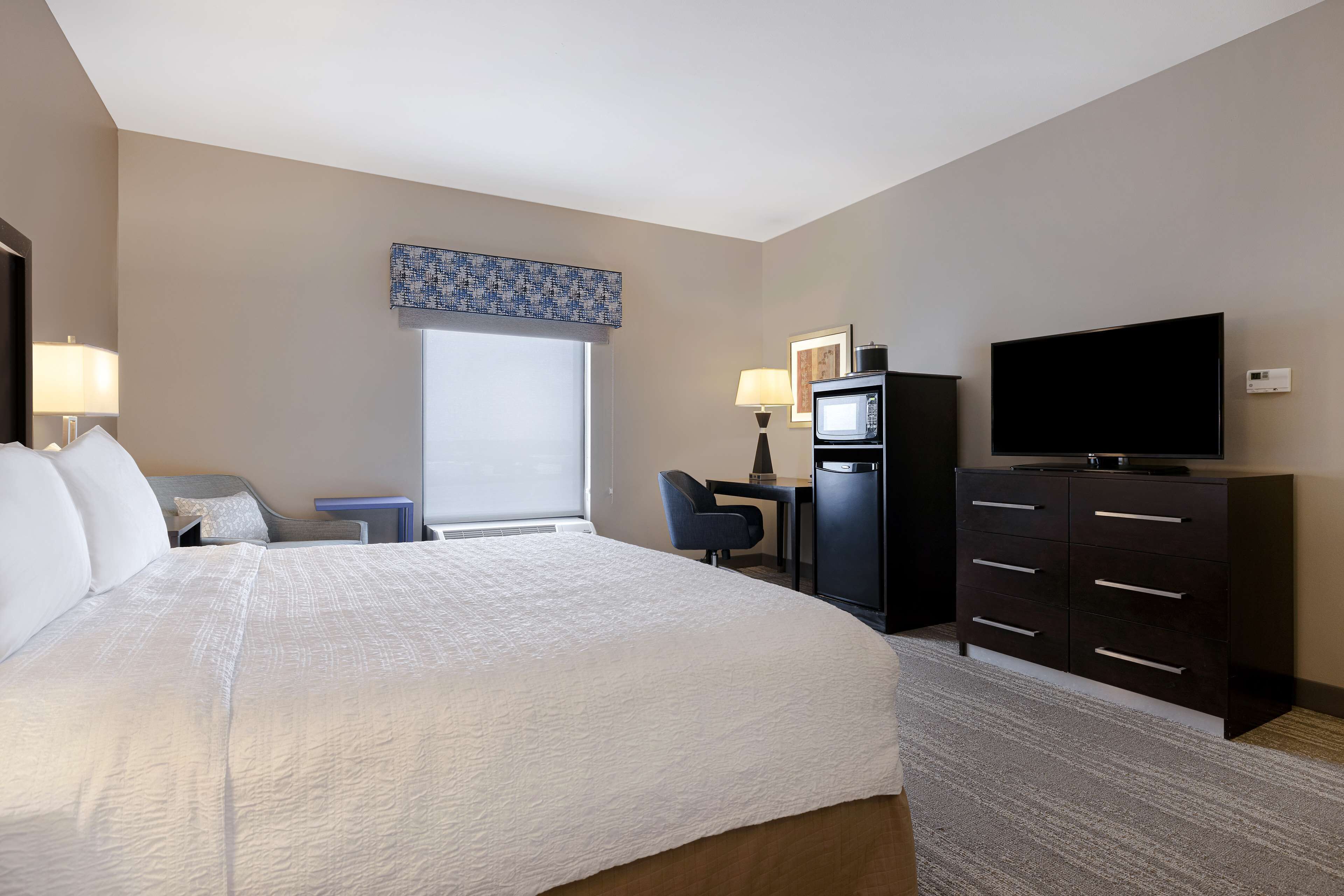 A hotel room with a large bed, a desk with a chair, a television, a dresser, and a window with blinds.