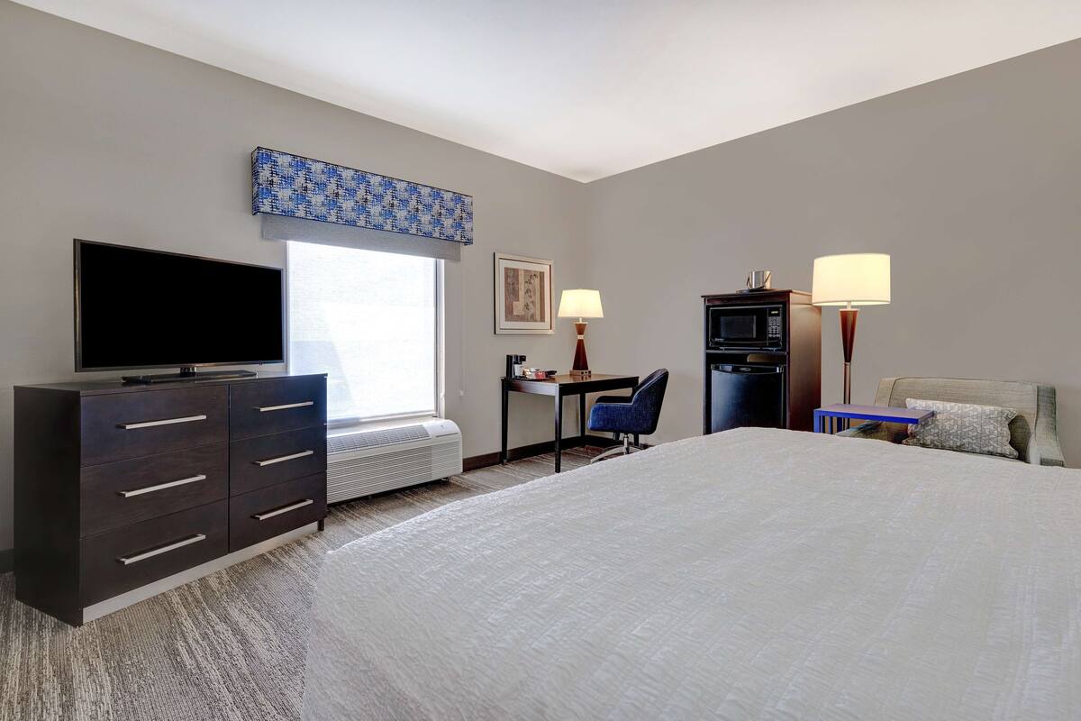 A hotel room with a large bed, a flat-screen TV, a desk with a chair, a microwave, and a small table with a lamp.
