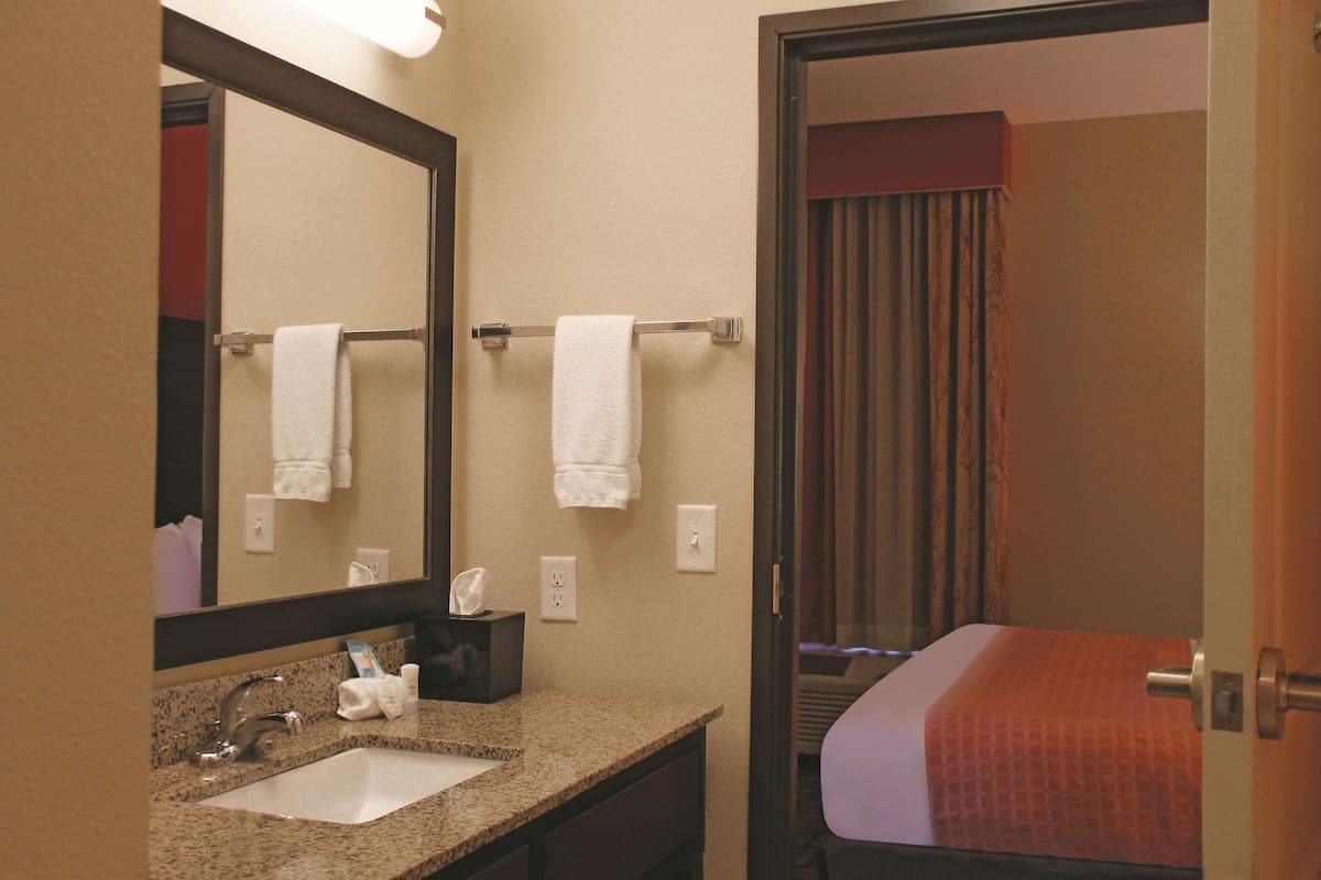 Une salle de bain d’hôtel avec un lavabo, un miroir, un porte-serviettes et un lit visible à travers une porte ouverte.