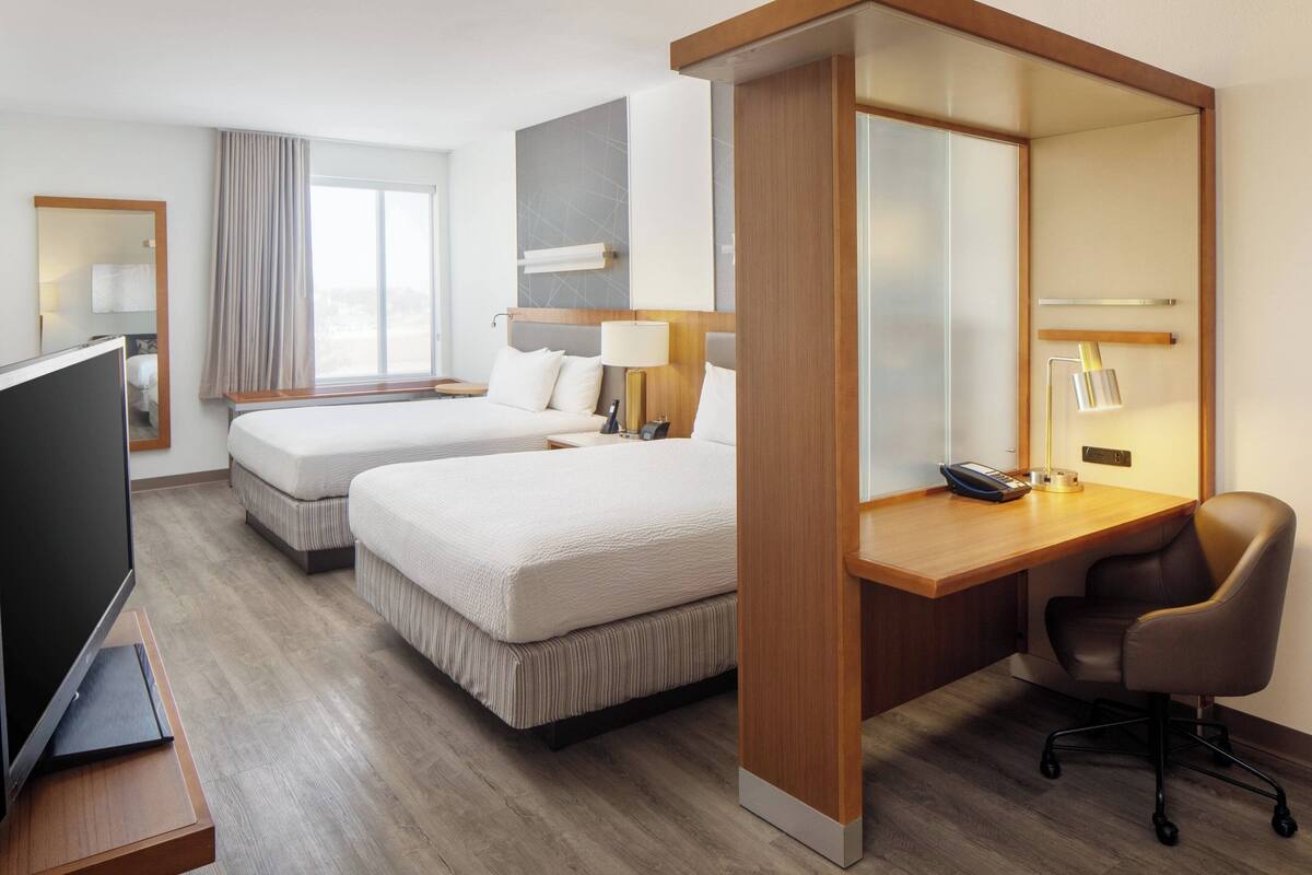 A hotel room with two beds, a desk, a chair, a TV, and a large window.