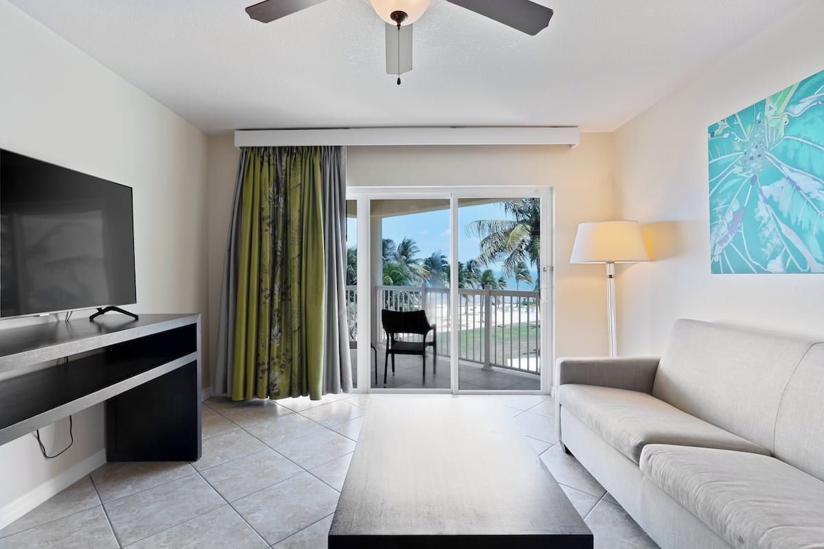A modern living room with a flat-screen TV, a sofa, a coffee table, and a balcony with a view of palm trees.