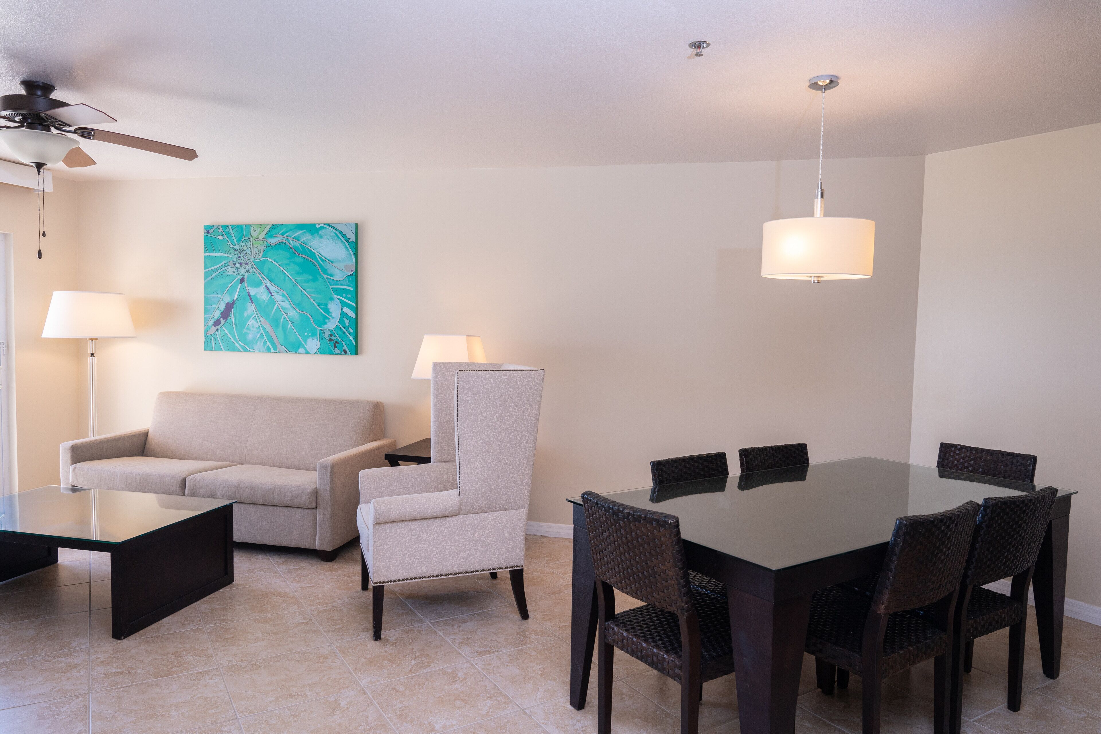Uma sala de estar moderna com sofá, poltrona, mesa de jantar e cadeiras. Há um ventilador de teto, um quadro na parede e um abajur sobre a mesa.