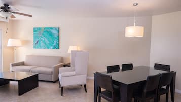 Uma sala de estar moderna com sofá, poltrona, mesa de jantar e cadeiras. Há um ventilador de teto, um quadro na parede e um abajur sobre a mesa.