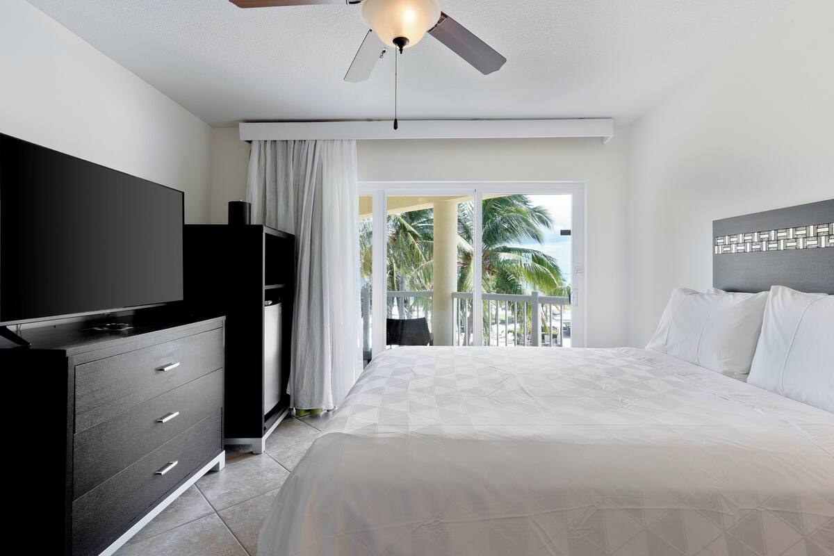 A modern hotel room with a bed, a TV, a ceiling fan, and a balcony with a view of palm trees.