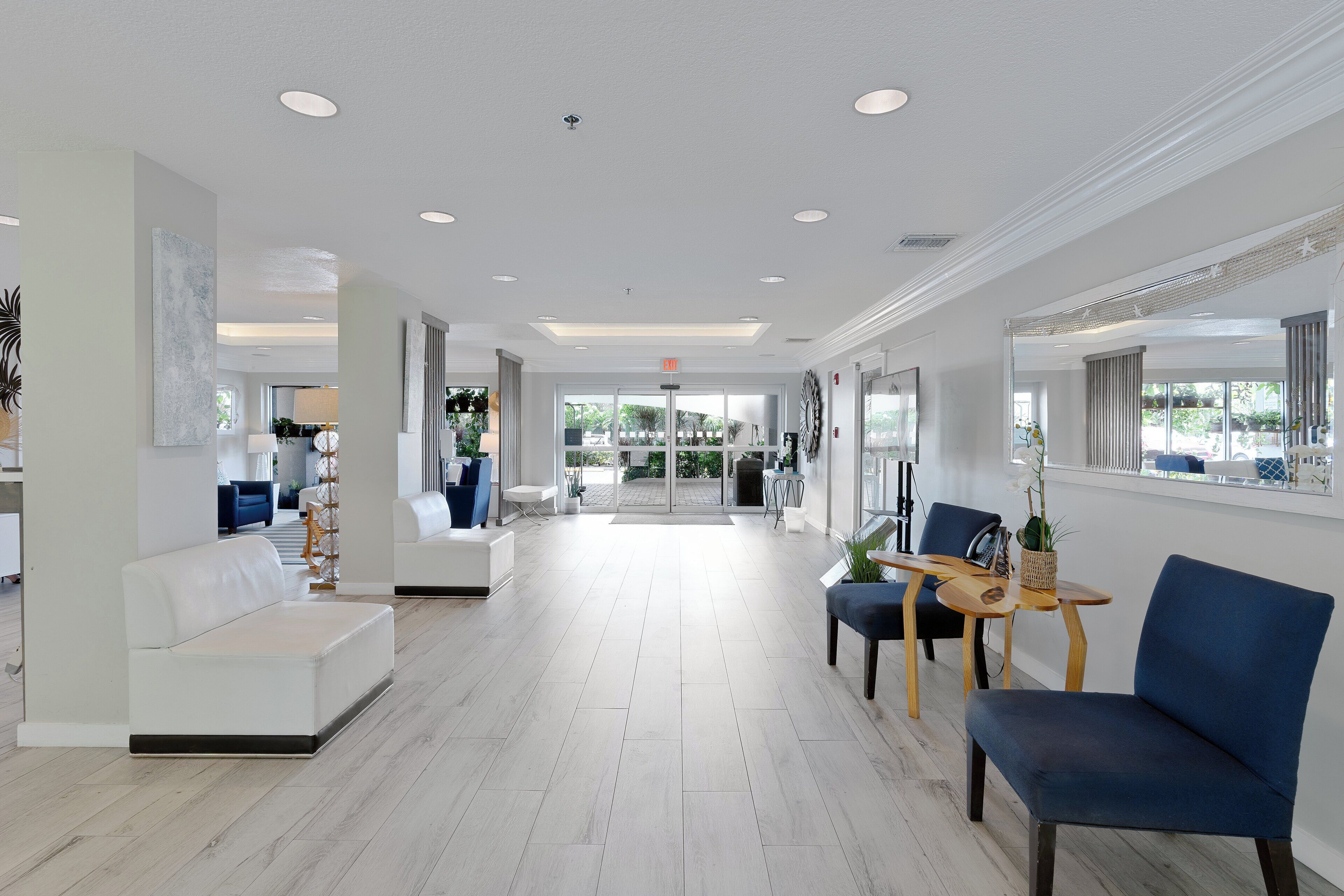 Un hall d’hôtel spacieux au design moderne, avec un espace salon central comprenant un canapé blanc et des fauteuils bleus, une table basse en bois et une grande fenêtre donnant sur l’extérieur.