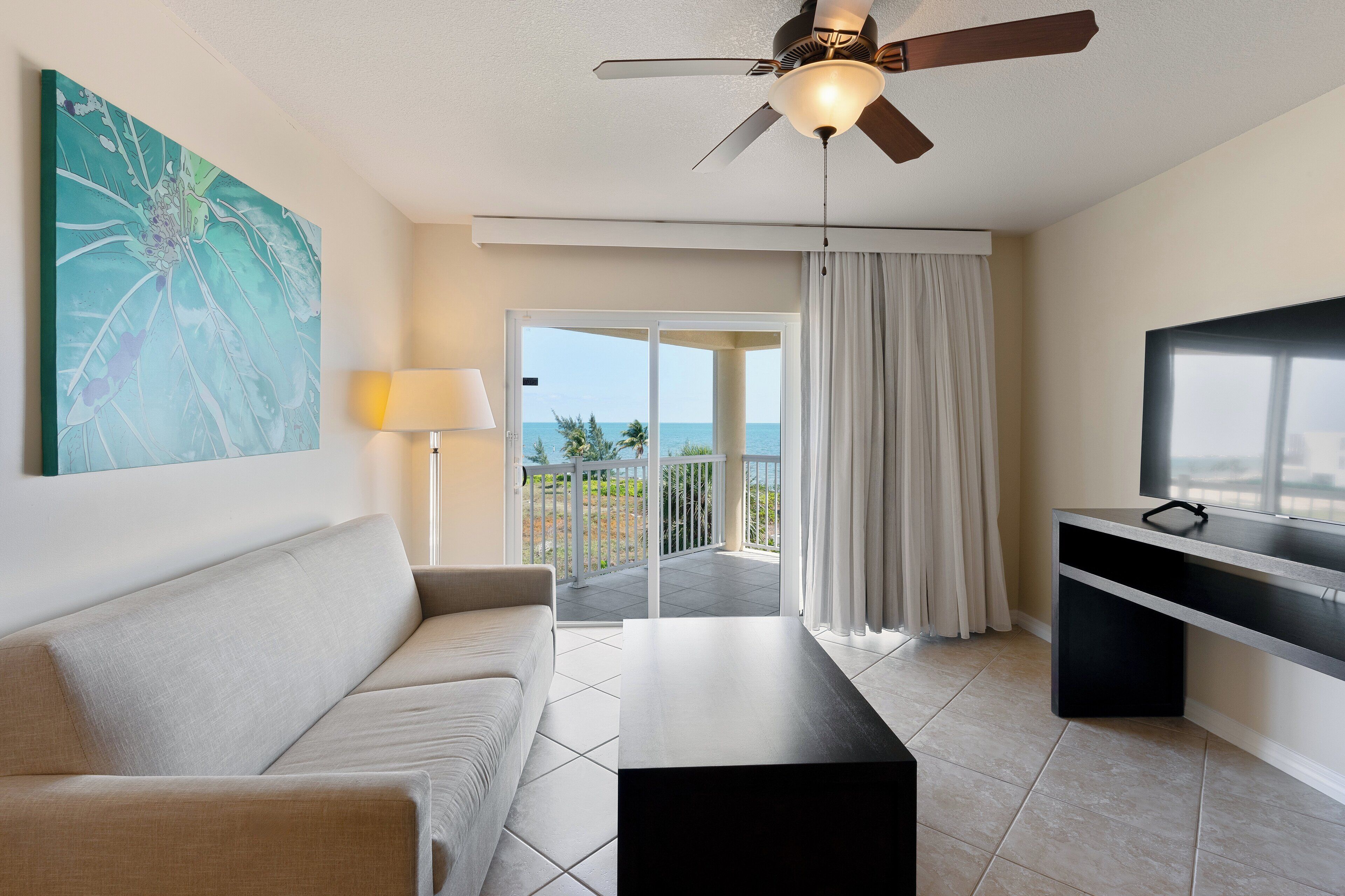 A hotel room with a sofa, a TV on a stand, a ceiling fan, a painting on the wall, and a view of the sea through the balcony doors.