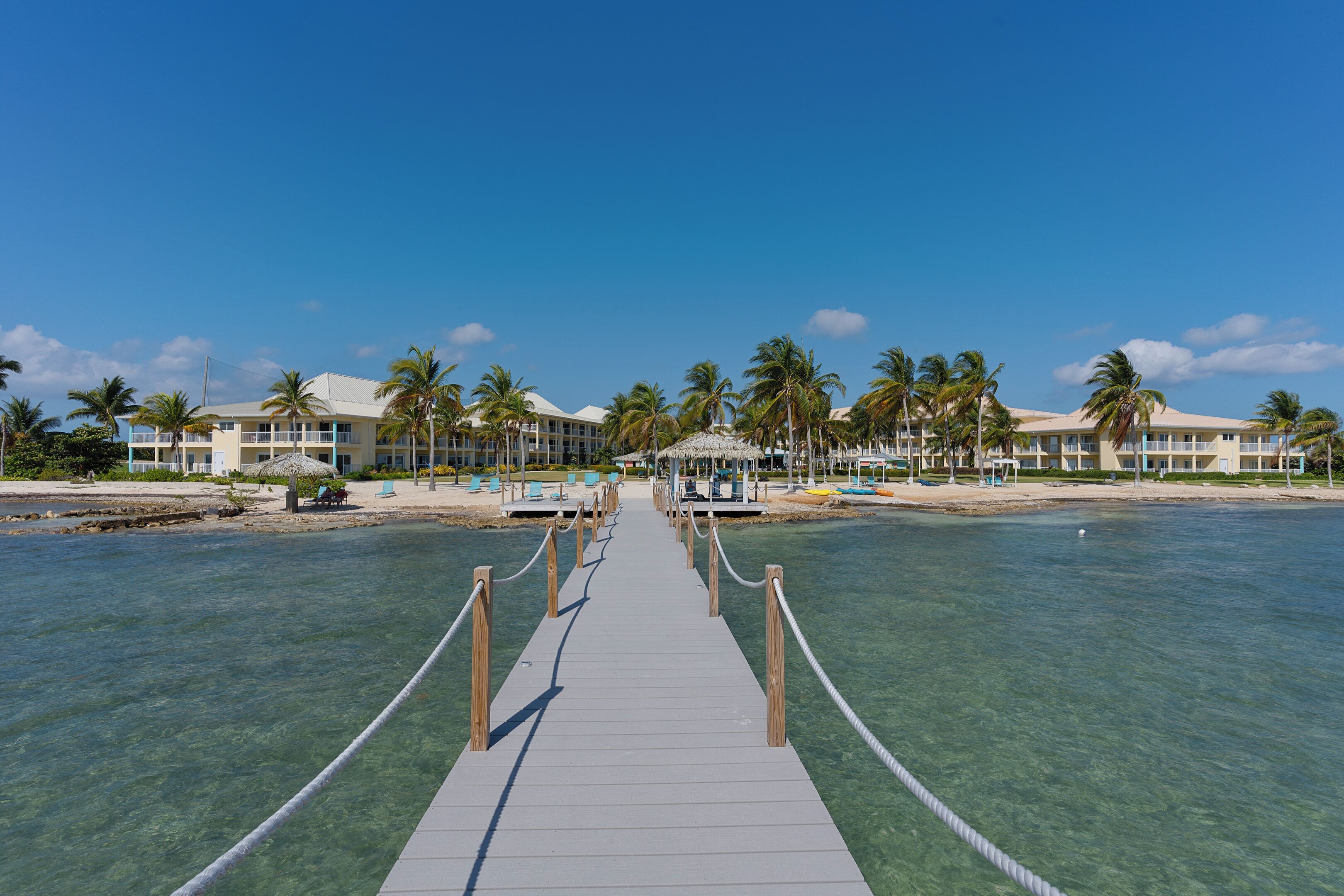 A clear water pathway leading to a resort with multiple buildings, palm trees, and a beach.