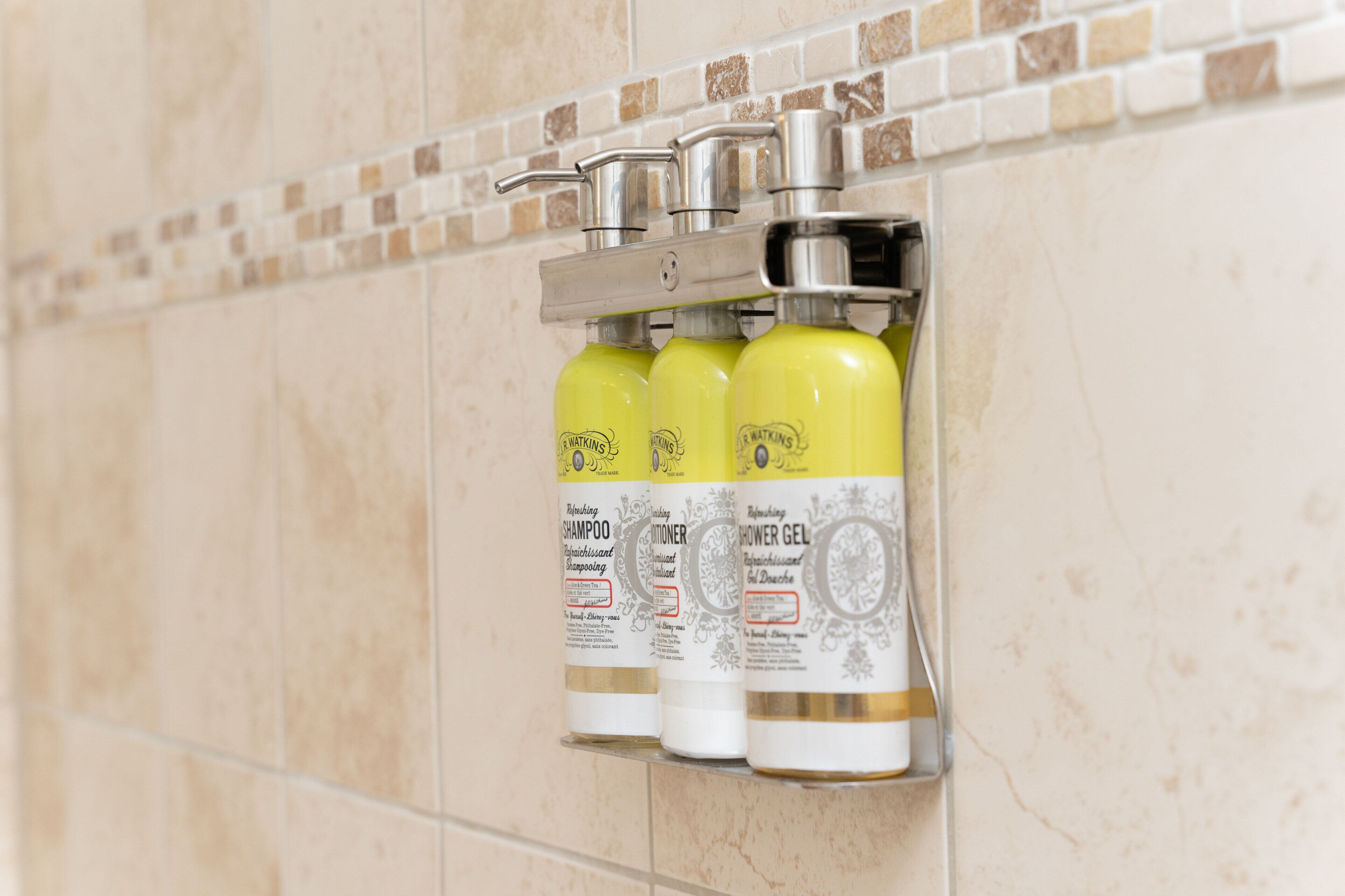 A bathroom with a tiled wall and a shelf holding three bottles of shampoo and conditioner.