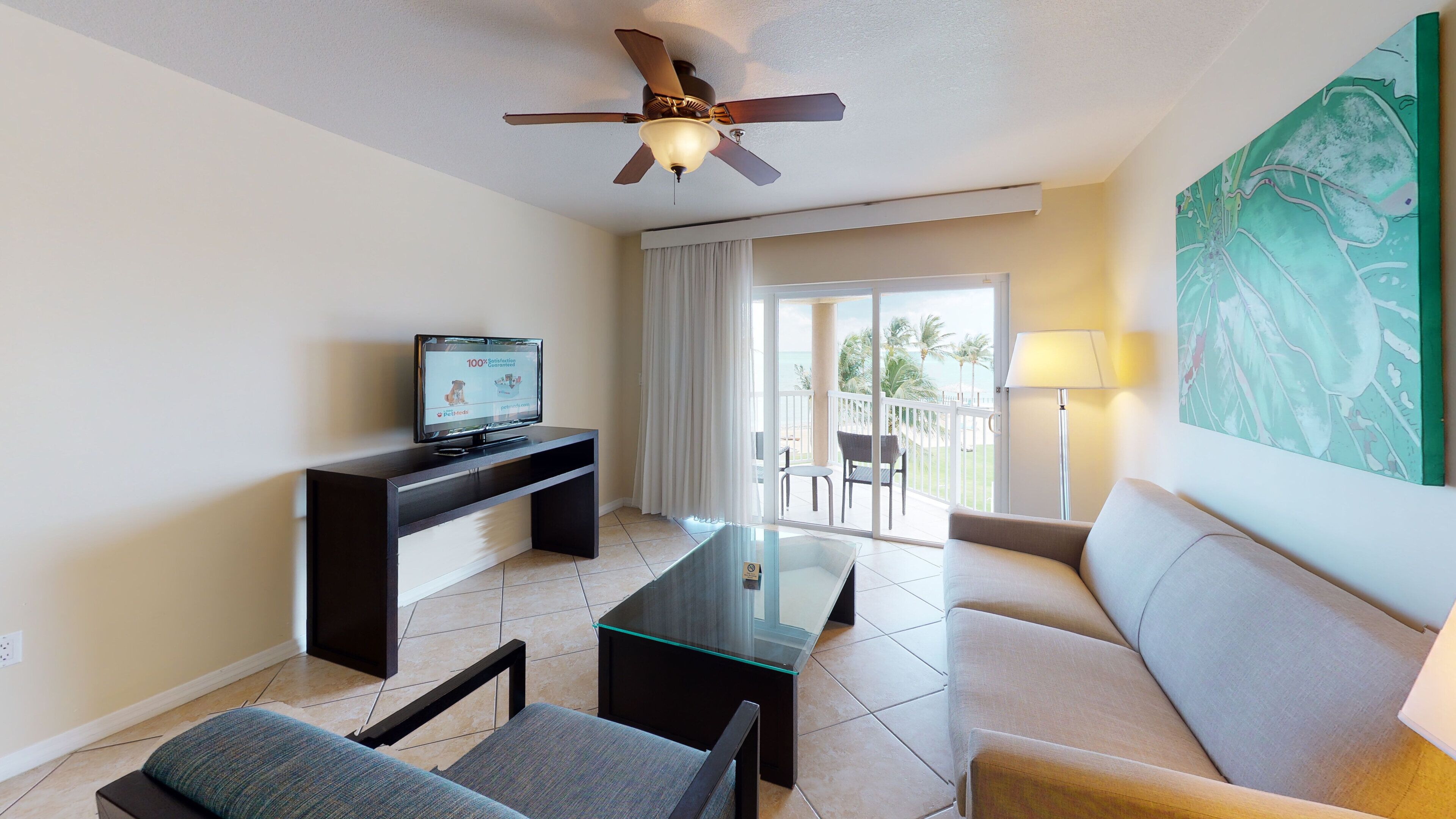 A modern hotel room with a flat-screen TV, a glass-top coffee table, a sofa, a ceiling fan, and a balcony with a view of the ocean.