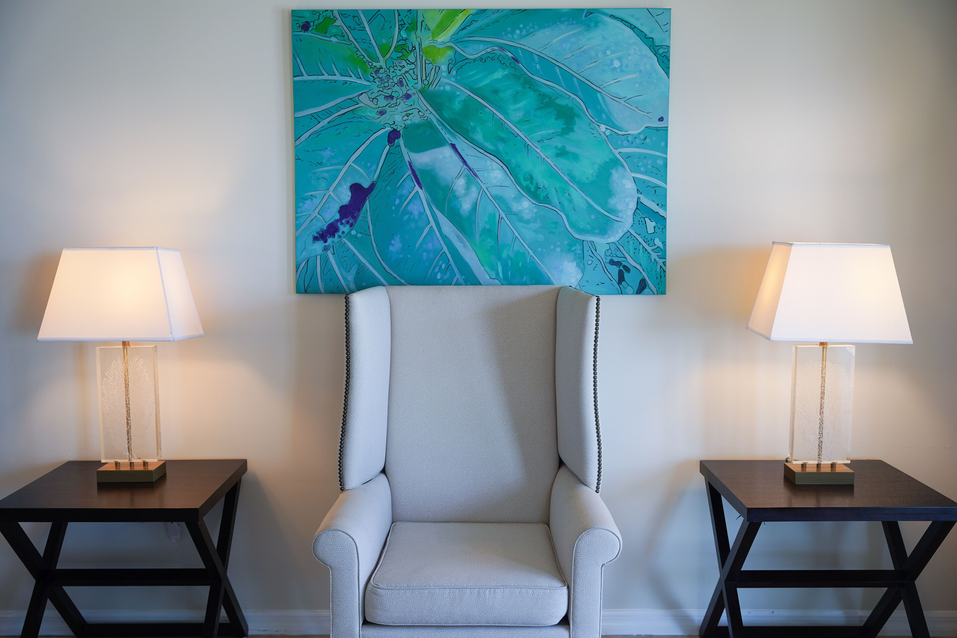 A modern room with a light grey armchair centered, flanked by two small tables with lamps. A large abstract painting adorns the wall above the armchair.