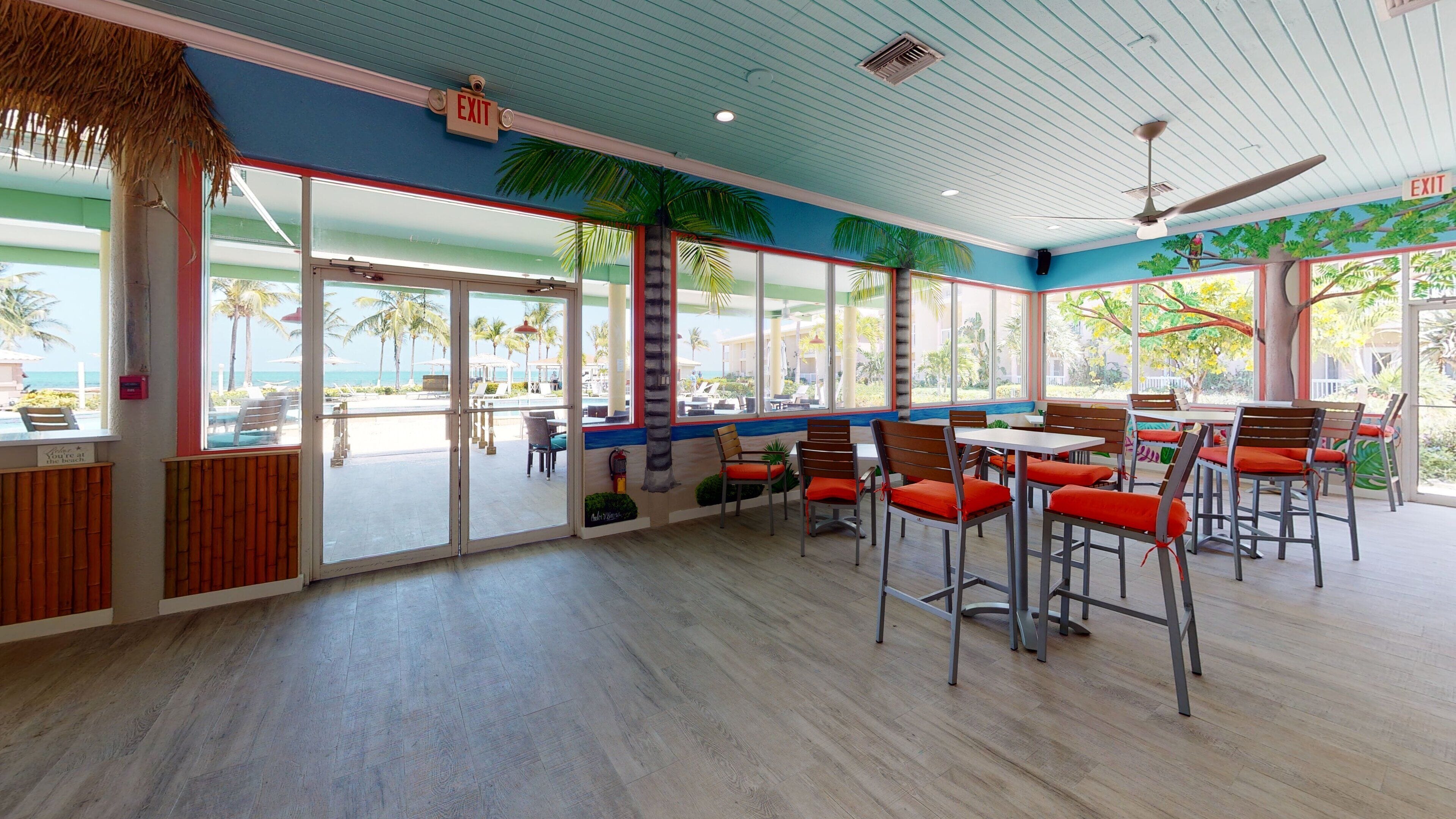 A restaurant with a view of the beach, featuring a tropical theme with palm trees and a blue ceiling.