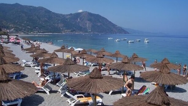 Ein Strand mit reetgedeckten Sonnenschirmen und Liegestühlen, einem klaren blauen Meer und fernen Bergen.