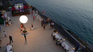 Ein Außenbereich für Veranstaltungen mit Tischen zum Essen, einer Bühne mit einer großen Leinwand und Blick auf das Meer.