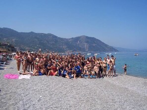 Un grupo de personas en una playa pedregosa con barcos en el agua y montañas al fondo.
