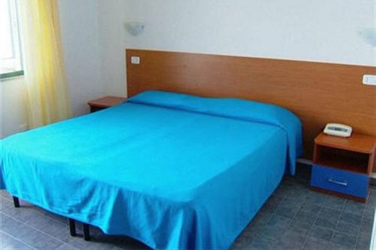 Ein Einzelbett mit blauer Bettdecke in einem Zimmer, das ein Holz-Kopfteil, einen Nachttisch mit Telefon und eine kleine Kommode hat.