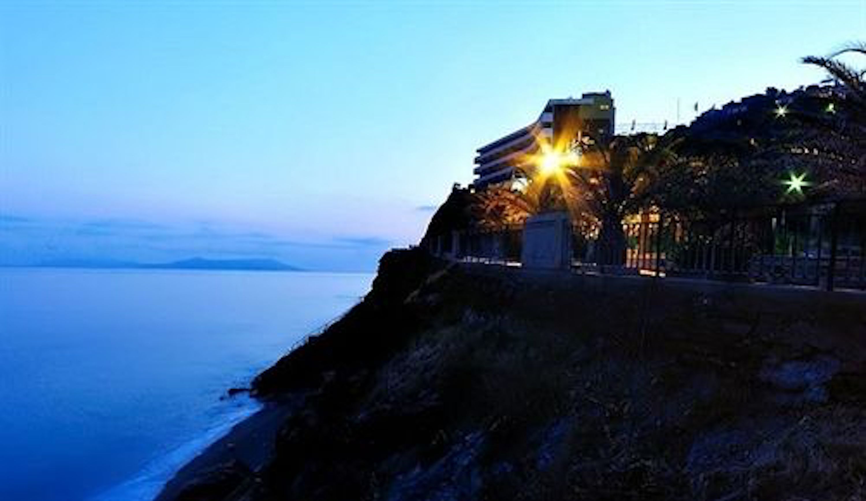 A coastal building with a balcony overlooking the sea, featuring a prominent staircase and a small garden area.