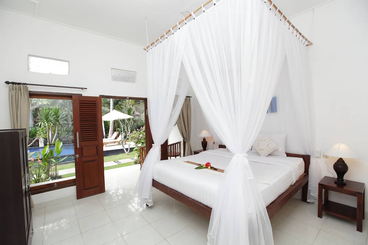 A bedroom with a bed, a nightstand, a lamp, a window, and a view of a pool and greenery.