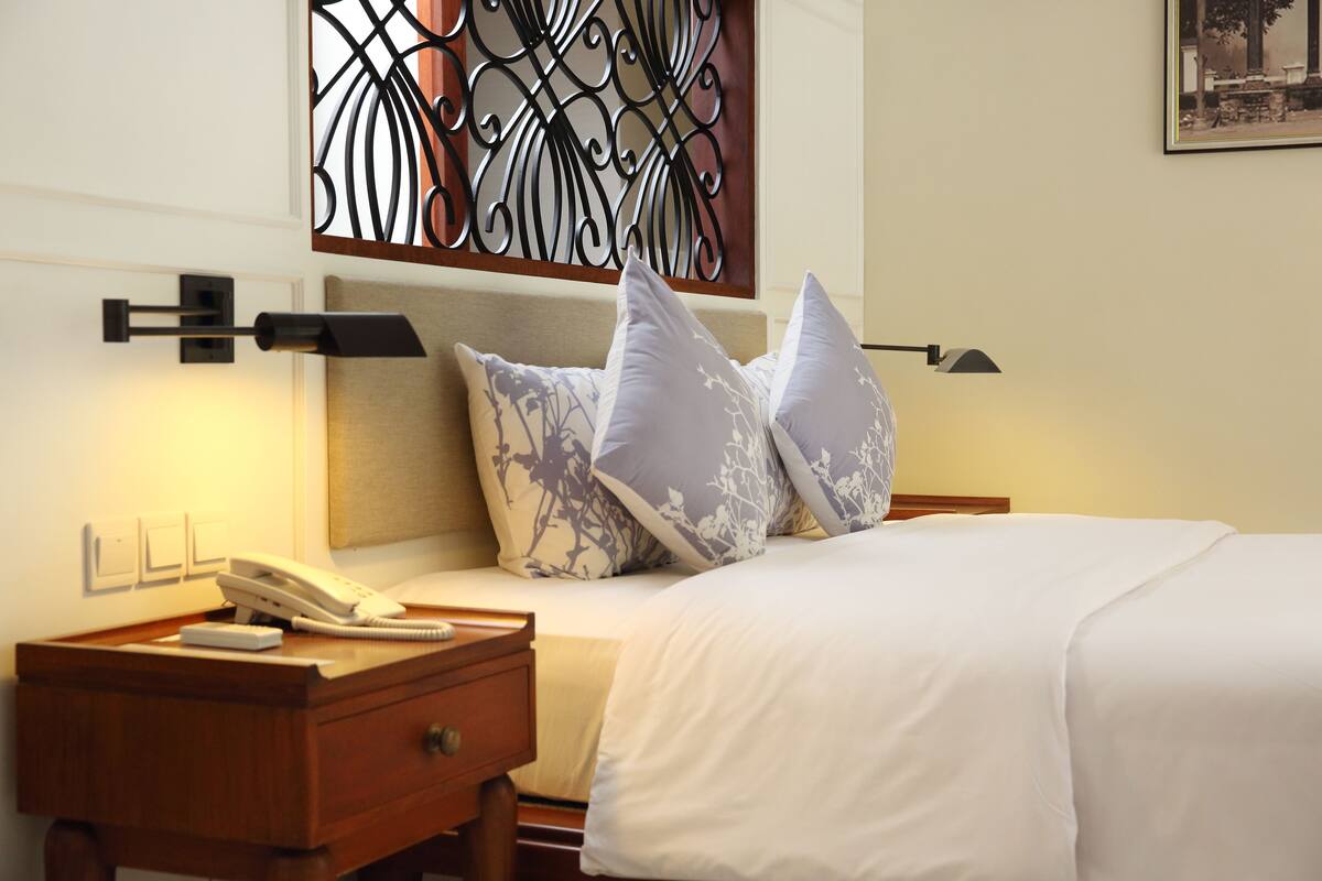 A neatly made bed with a white comforter and patterned pillows, a bedside table with a telephone, and a decorative wall piece above the bed.