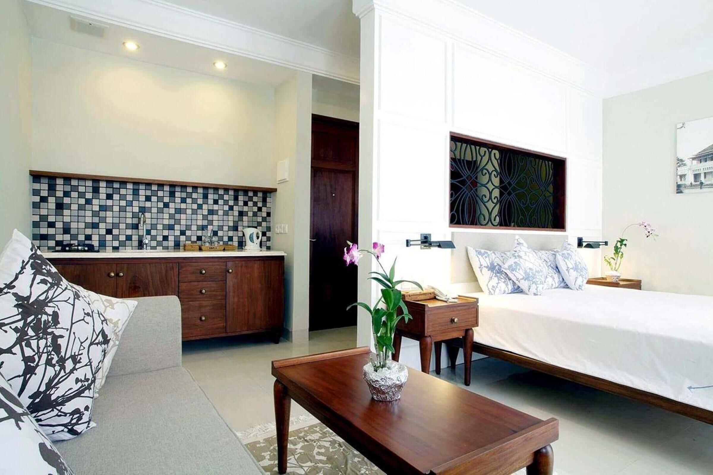 A modern hotel room with a bed, a wooden nightstand, a desk, a sofa, and a kitchen area with a tiled backsplash.
