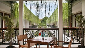 A balcony with a table and chairs, surrounded by potted plants and a view of a pool and greenery. - Seminyak Lagoon All Suites Hotel (Keroboken Kelod)