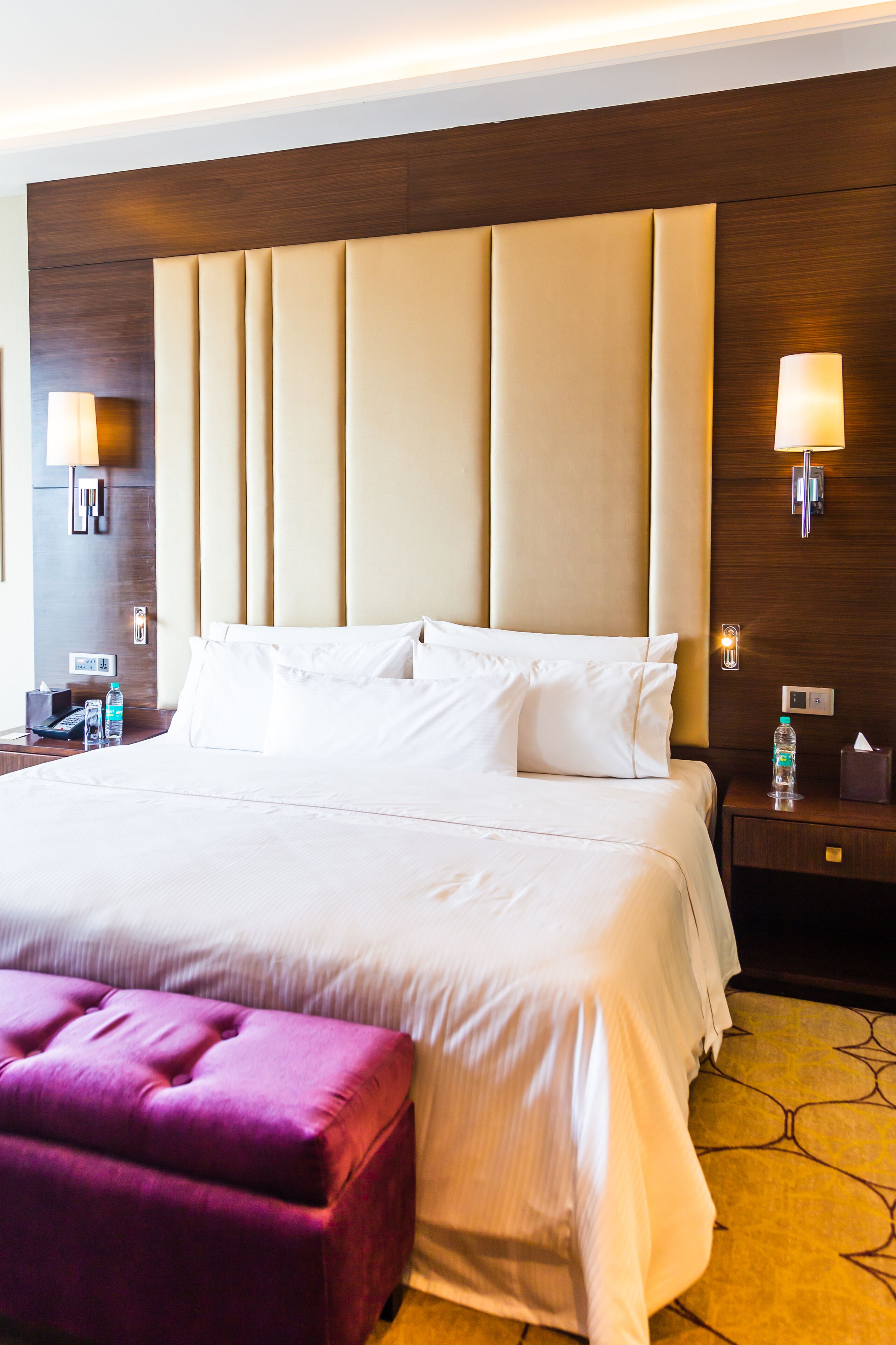 A hotel room with a large bed, a wooden headboard, a nightstand with a lamp, a water bottle, and a purple ottoman.