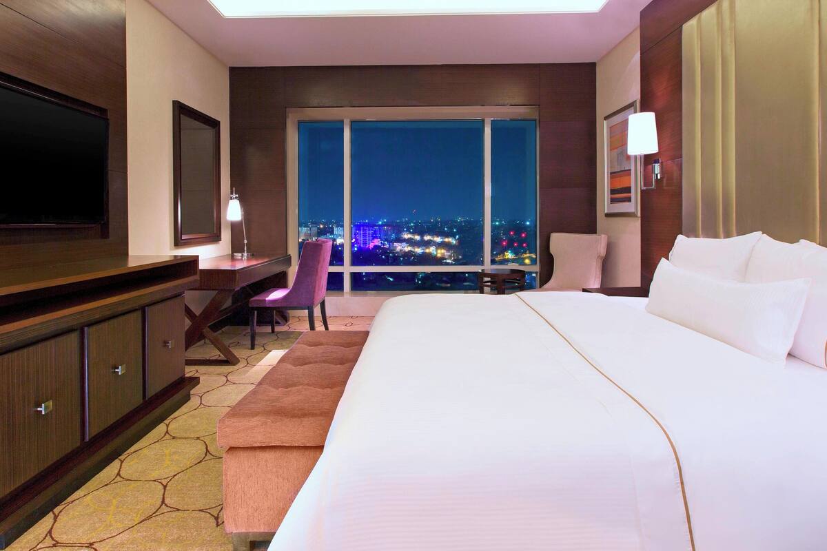A hotel room with a large bed, a TV, a desk, and a chair. There is a view of the city at night through the windows.