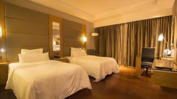 A hotel room with two beds, a desk, a chair, a lamp, and a large window with curtains.