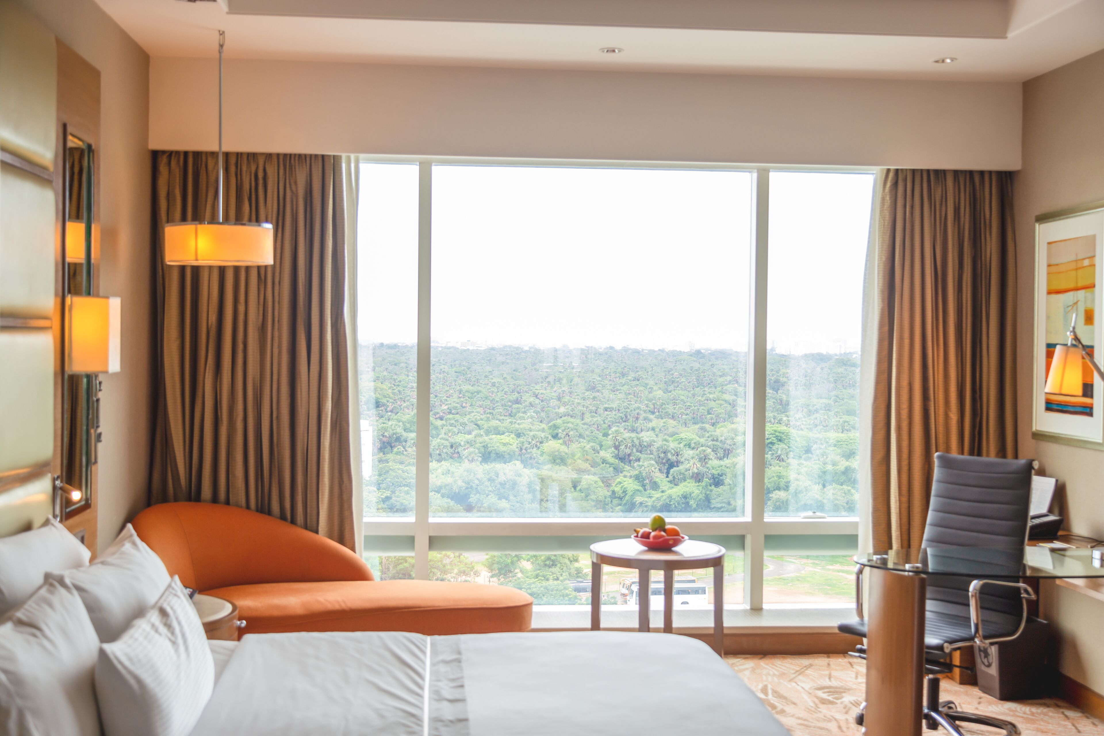 Une chambre d’hôtel avec un lit, un fauteuil orange, une petite table ronde sur laquelle se trouve un panier de fruits, un fauteuil de bureau noir et une grande fenêtre donnant sur une zone boisée.