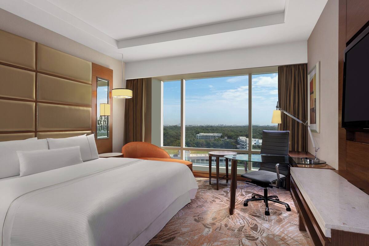 A hotel room with a large bed, a desk with a chair, a small table, and a large window offering a city view.