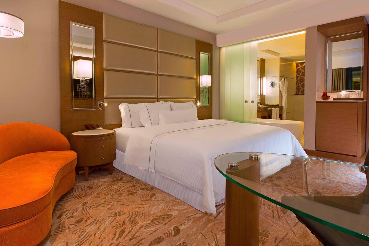 A hotel room with a large bed, a glass table, a sofa, a nightstand, and a bathroom visible through an open door.
