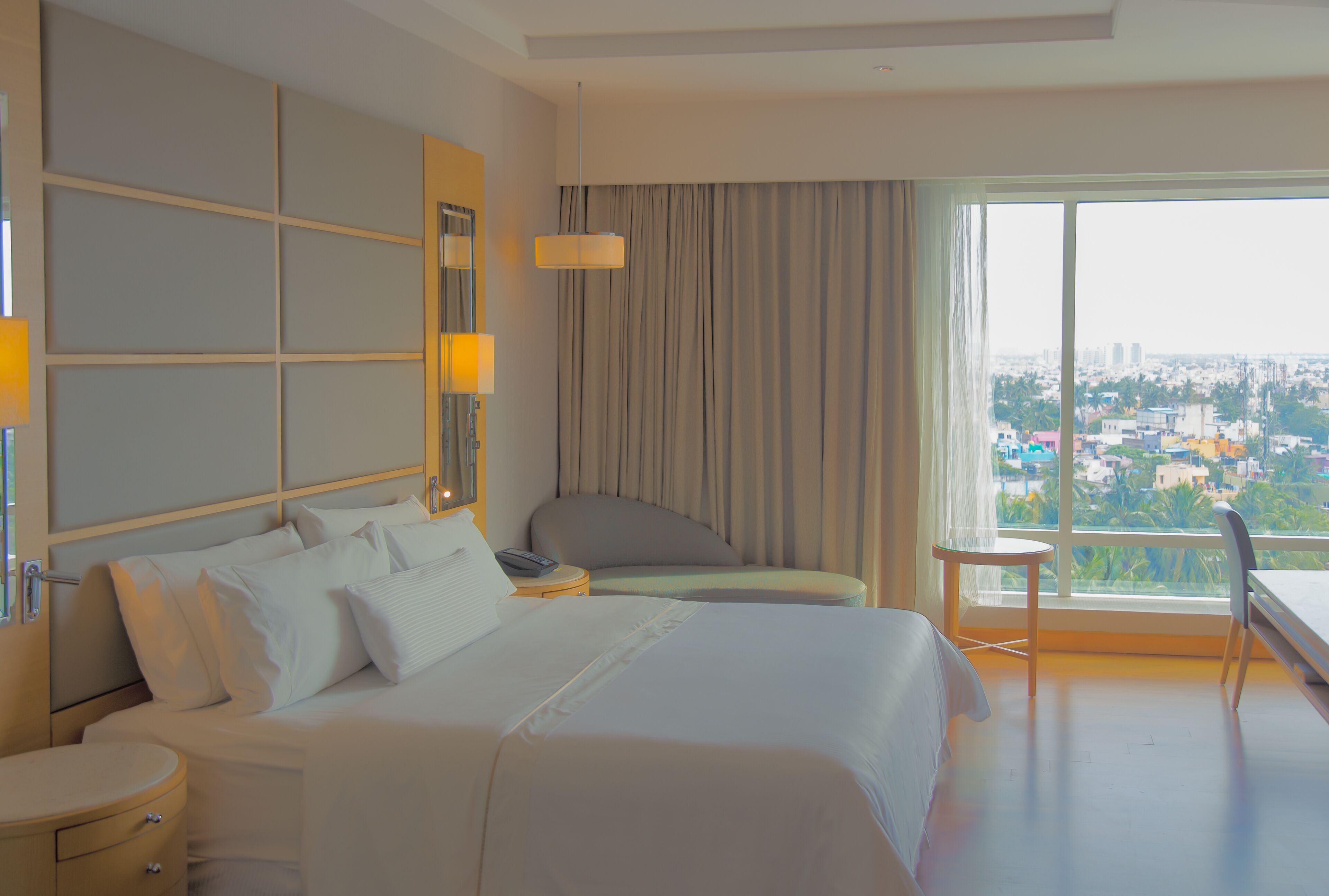 Habitación de hotel con una cama grande, una mesita de noche, una silla, una pequeña mesa y vistas de la ciudad a través de un ventanal amplio.