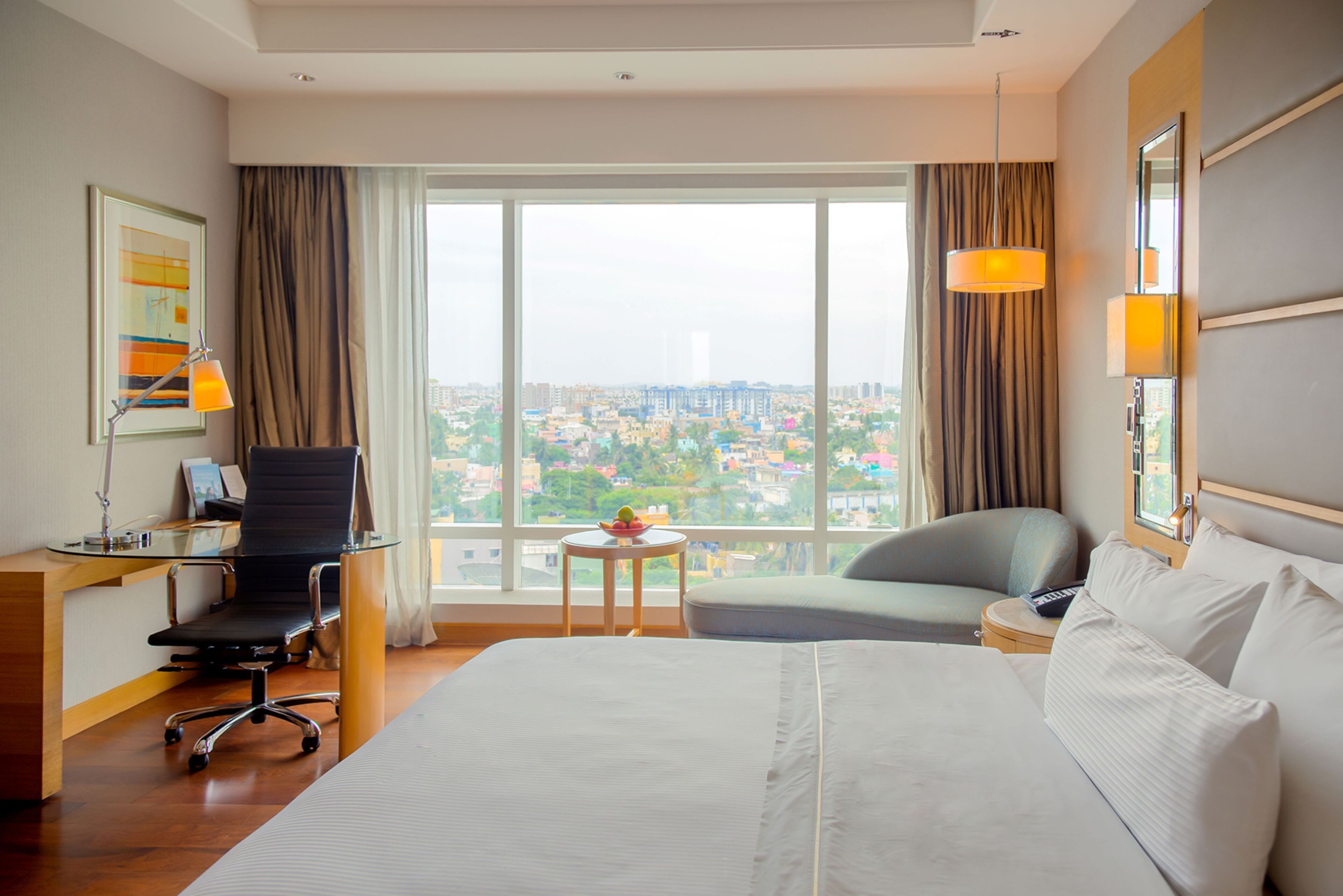 Une chambre d’hôtel avec un grand lit, un bureau avec une chaise, une petite table sur laquelle se trouve un saladier rempli de fruits, et une vue sur le paysage urbain à travers de grandes fenêtres.