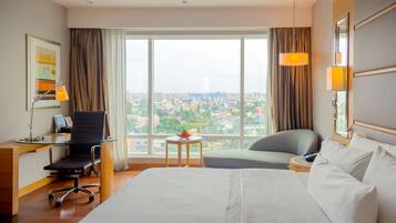 Une chambre d’hôtel avec un grand lit, un bureau avec une chaise, une petite table sur laquelle se trouve un saladier rempli de fruits, et une vue sur le paysage urbain à travers de grandes fenêtres.