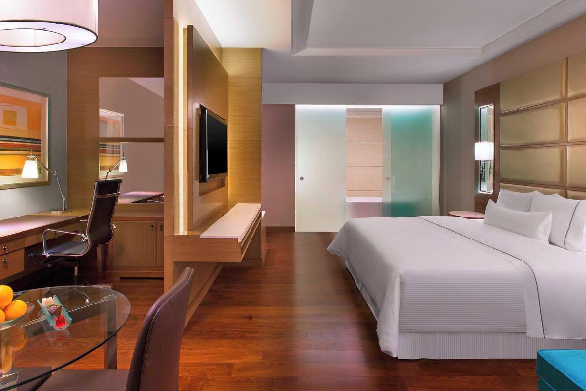 A modern hotel room with a bed, a desk with a chair, a TV, and a glass table with fruits.