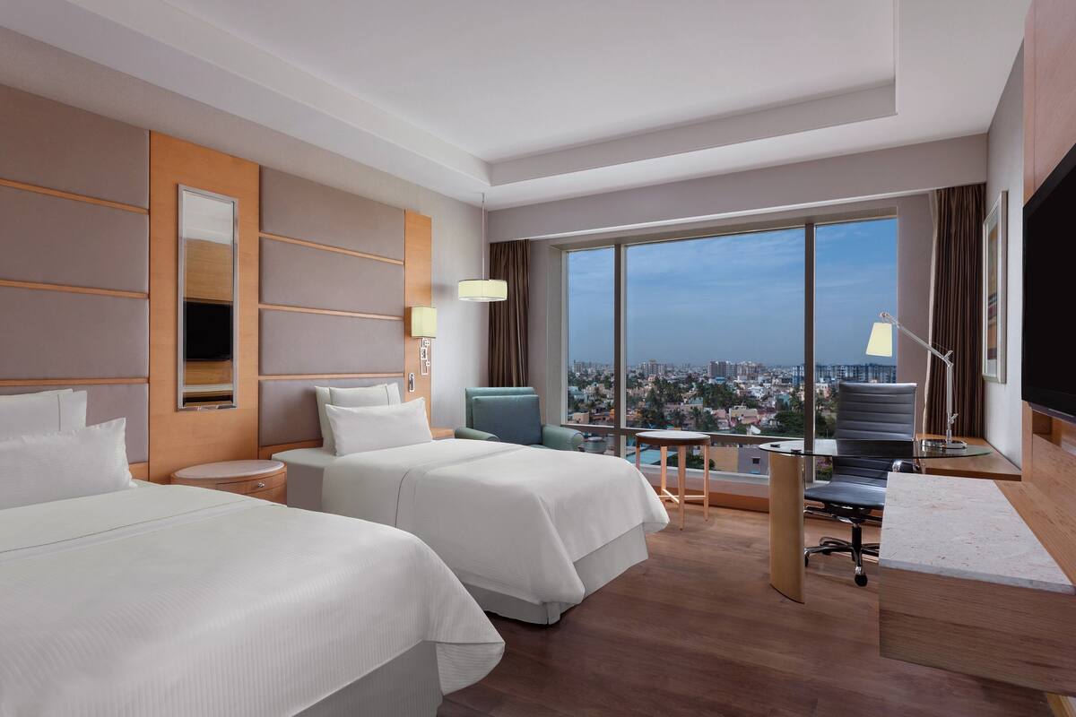 A hotel room with two beds, a desk, a chair, a TV, and a large window with a city view.