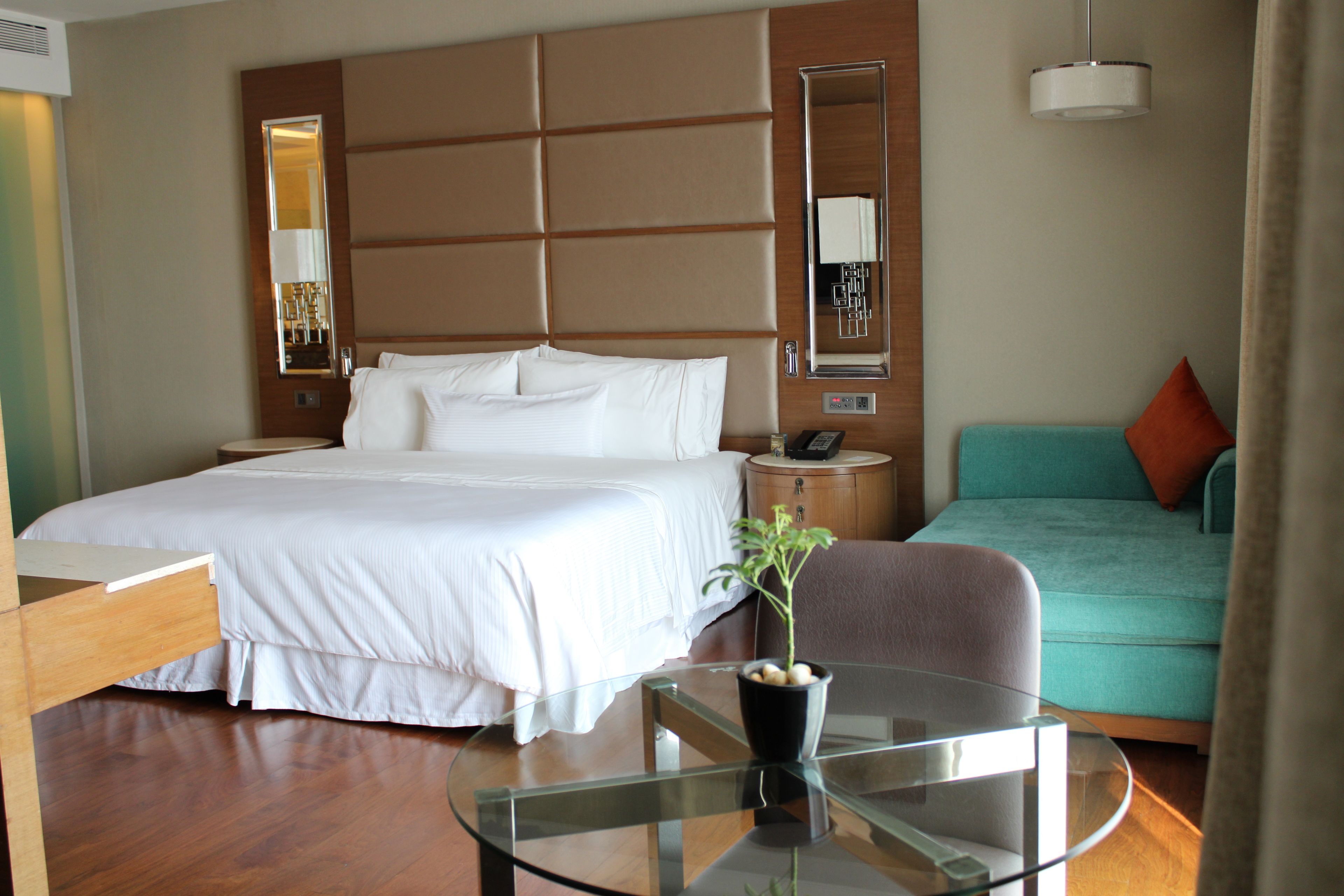 A hotel room with a large bed, a glass table, a small plant, a sofa, and a mirror.