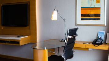 A modern hotel room with a flat-screen TV, a desk with a telephone, a glass table with fruit, and a framed abstract painting on the wall.