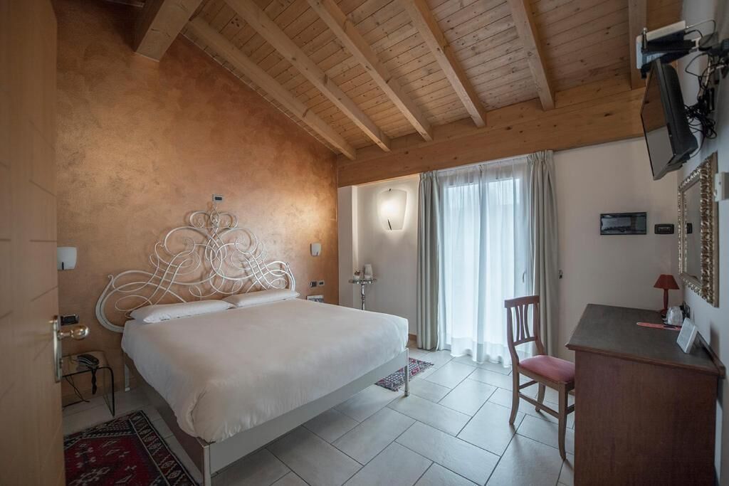 Ein Schlafzimmer mit Holzbalkendecke, einem weißen Bett mit einem besonderen Kopfteil, einem Schreibtisch mit Stuhl und einem großen Fenster mit Vorhängen.