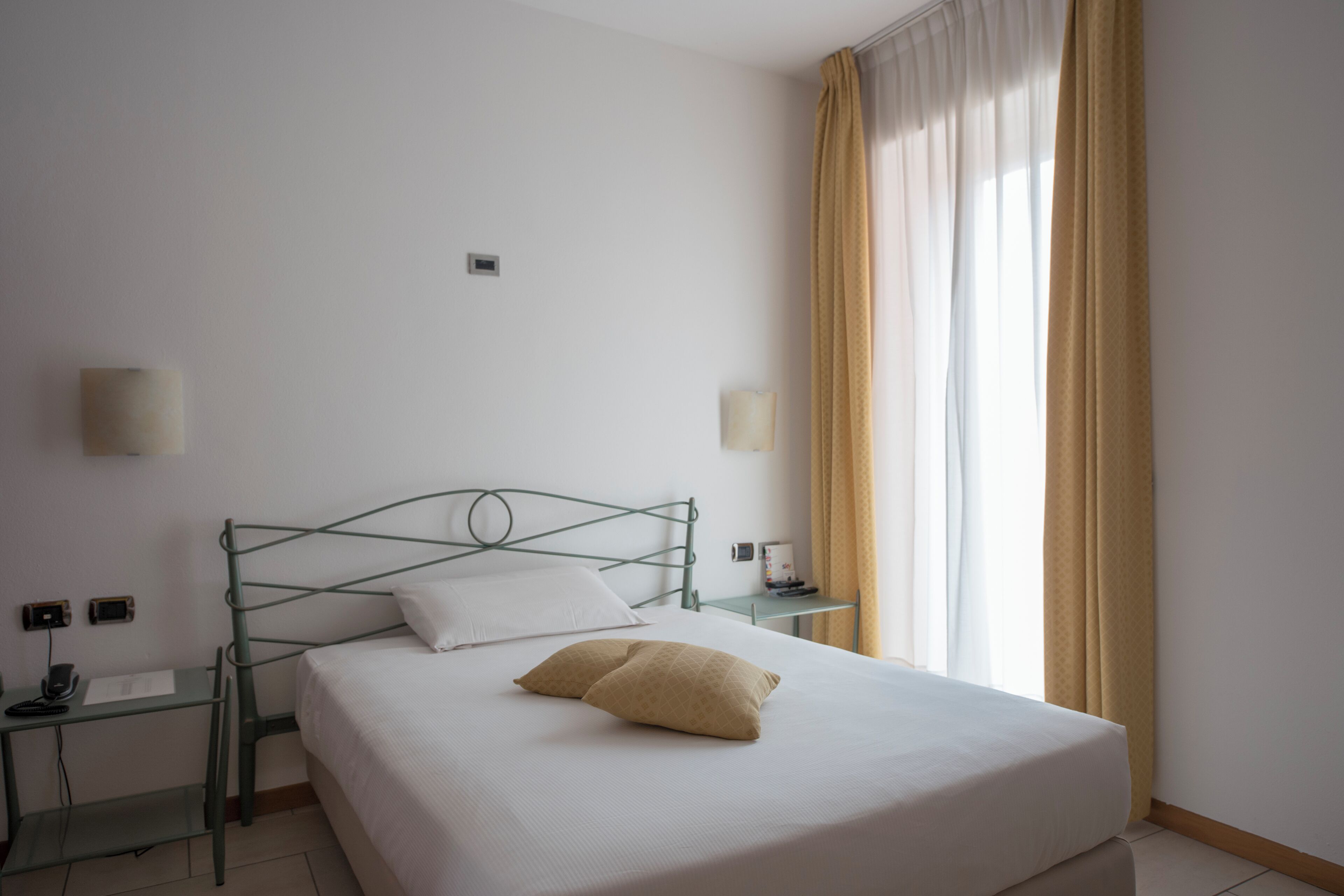 A neatly made bed with a single pillow, a bedside table with a lamp, and a window with curtains.