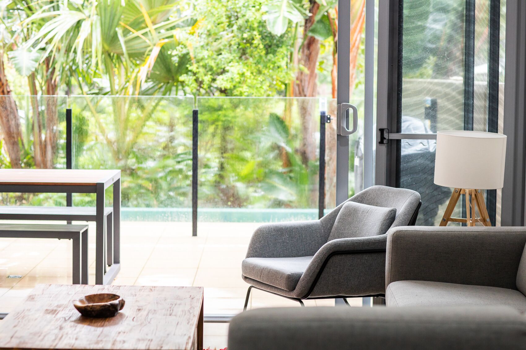 A modern living room with a sofa, armchairs, and a wooden coffee table. A glass door leads to a balcony with a view of lush greenery.
