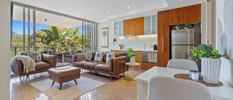 Una sala de estar moderna con un ventanal grande, un conjunto de sofás, una mesa de comedor y una cocina con armarios de madera y un refrigerador.