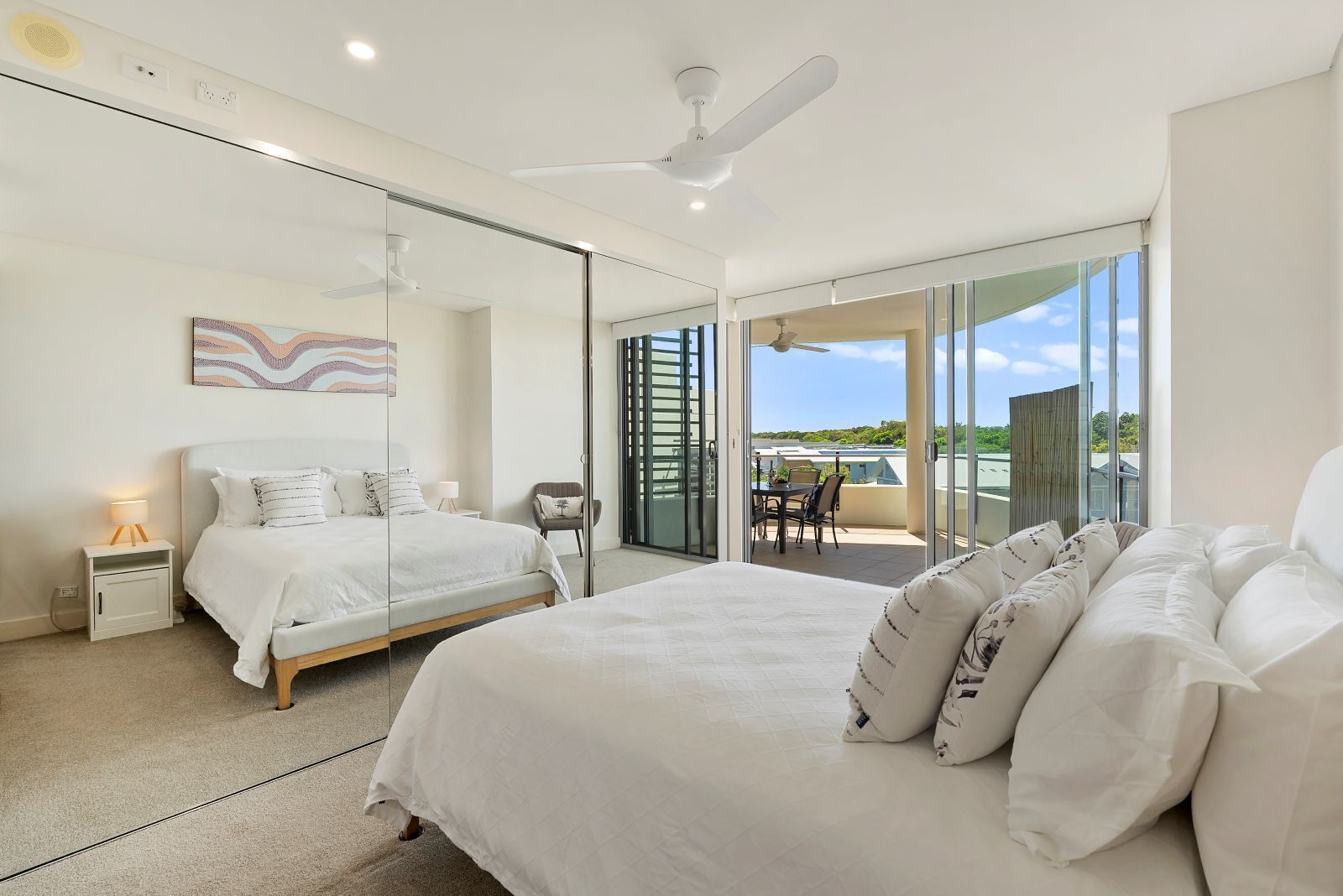 A modern bedroom with a large bed, a smaller bed, a chair, a table, and a balcony with a view.