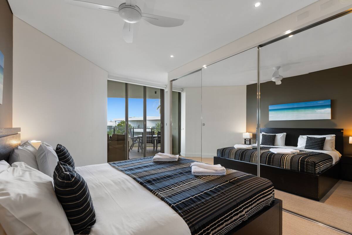 Een moderne hotelkamer met een groot bed, een klein bed, een plafondventilator, een schuifdeur naar een balkon en een schilderij aan de muur.
